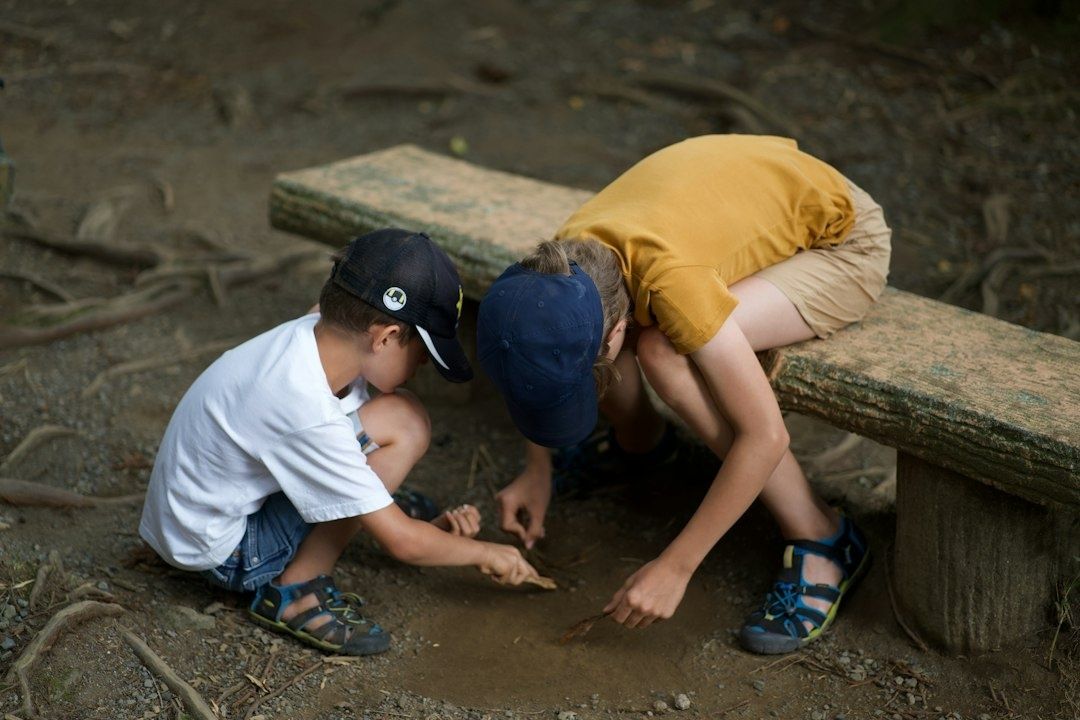 Three children investigate the ground outdoors beside a stone bench